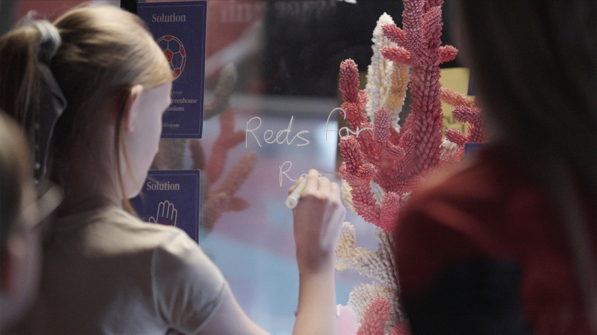 Young participant writing 'Reds for Red' on the glass case of the coral sculpture